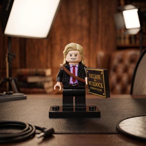 May include: A Lego minifigure with blonde hair, wearing a black suit jacket over a light purple shirt with a red patterned tie. A brown strap crosses the chest. The figure holds a black rectangular sign with gold text reading Nelson and Murdock Attorneys at Law.