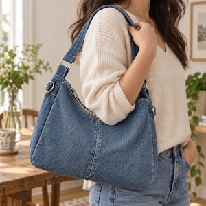 May include: A medium blue denim shoulder bag featuring a structured rectangular shape, a top zipper closure, and silver-toned metal hardware connecting the strap to the body. The bag is crafted from durable denim fabric with visible stitching details.