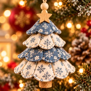 May include: A hanging Christmas tree ornament crafted from layered fabric petals in alternating blue and cream colors, featuring a white snowflake pattern. The ornament is topped with a small wooden star and a wooden bead, suspended by a twine loop.
