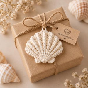 Puede incluir: Una caja de regalo cuadrada envuelta en papel marrón, atada con cordel marrón y adornada con un adorno de concha marina de ganchillo color crema. Una pequeña etiqueta rectangular unida al cordel muestra el texto HealAndHookCo y un icono.