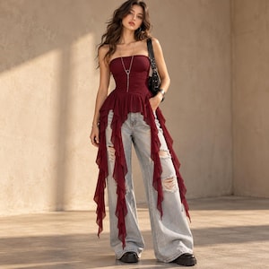 May include: A burgundy strapless top featuring a fitted bodice and long, cascading ruffled side panels, paired with light-wash, wide-leg distressed denim jeans. The top includes a thin silver-toned necklace with a circular pendant.