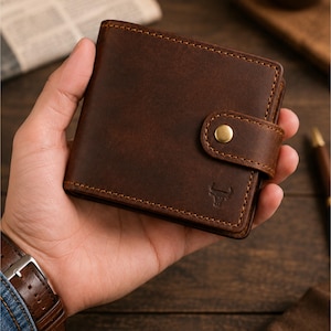 Puede incluir: Una cartera bifold de cuero marrón oscuro con una lengüeta de cierre de botón a presión y un pequeño logotipo de toro en relieve en la parte delantera. La cartera tiene costuras visibles a lo largo de los bordes y un acabado mate liso.