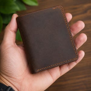May include: A dark brown leather wallet featuring a smooth, matte finish and visible orange stitching along the edges. The rectangular wallet is folded and displays a clean, minimalist design with a consistent texture across the front surface.