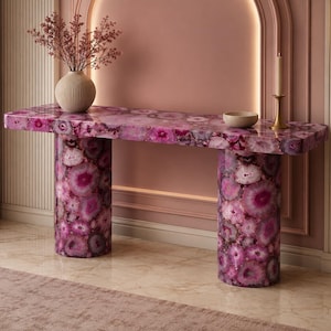 May include: A console table constructed from polished pink agate stone, featuring a rectangular top supported by two cylindrical columns. The surface displays a natural, multi-tonal pink and white crystalline pattern throughout the entire structure.