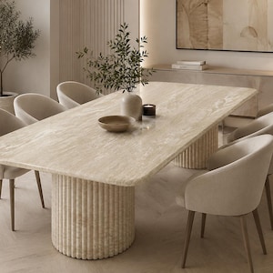 Puede incluir: Una mesa de comedor rectangular con tablero de piedra travertino beige claro y base cilíndrica estriada, rodeada por ocho sillas tapizadas en tela crema claro con patas finas marrones, con un cuenco y un jarrón sobre la superficie.
