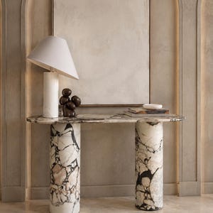 Pode incluir: Uma mesa consola com um tampo oval alongado e duas bases cilíndricas, feita de mármore branco com veios pretos e cinzentos proeminentes. Sobre a mesa encontram-se um candeeiro branco, uma escultura abstrata castanha e livros empilhados.