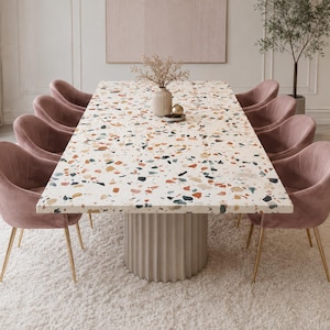 May include: A rectangular dining table with a white terrazzo top featuring multicolored stone chips and a fluted cylindrical base, surrounded by eight upholstered pink velvet chairs with slender gold-toned metal legs.