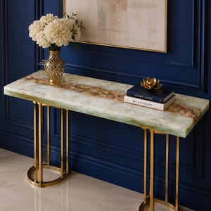 May include: A rectangular console table with a polished green and brown stone top and two gold-toned metal bases. A gold lattice vase with white flowers, two stacked books titled Luxury Living and Art & Interiors, and a gold lotus ornament sit on top.