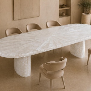 May include: An oval dining table with a white, sunburst-patterned stone surface and two matching curved pedestal bases, accompanied by six beige upholstered chairs with rounded backrests and slender legs arranged around the table.