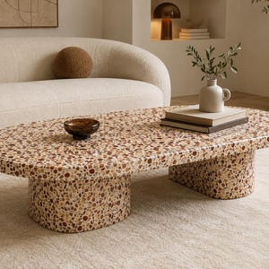 Könnte beinhalten: Ein ovaler Couchtisch mit einer gesprenkelten Terrazzo-Oberfläche in Braun-, Beige- und Cremetönen. Der Tisch wird von zwei breiten, zylindrischen Sockeln getragen, die durchgehend das gleiche Terrazzo-Muster aufweisen.