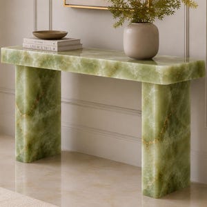 Puede incluir: Una mesa consola rectangular hecha de piedra ónix verde claro con vetas naturales e inclusiones doradas. La mesa presenta una superficie gruesa y plana sostenida por dos patas rectangulares anchas y sólidas, con un acabado pulido y suave.