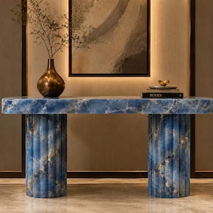 May include: A blue stone console table with a smooth rectangular top and two fluted cylindrical bases. A dark metallic vase with thin branches and a stack of books with the word RODIN on the spine are placed on the surface of the table.