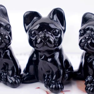 May include: Three small, glossy black figurines shaped like French Bulldog puppies. Each sculpture features upright ears, a short snout, and a smooth, reflective surface. The figures are arranged in a row, showcasing a consistent dark colour.