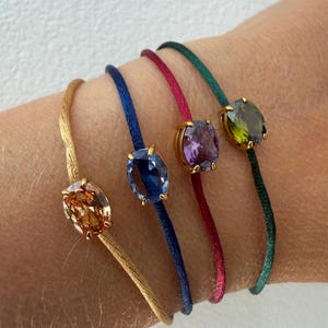 Pode incluir: Quatro pulseiras com pedras preciosas facetadas de formato oval, engastadas em garras de metal dourado. As bandas são feitas de cordão fino e liso nas cores dourado, azul, magenta e verde escuro, cada uma com uma pedra de tom similar.