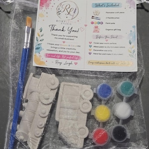 May include: A craft kit in a sheer organza bag containing two unpainted train-shaped plaster figures, a blue-handled paintbrush, and a strip of six small paint pots in blue, yellow, red, green, white, and black, with an instructional card.
