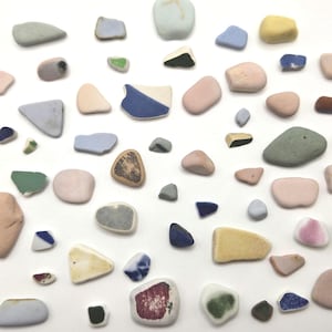 May include: A collection of assorted sea glass pieces in various shapes and sizes, featuring a palette of soft pastel pinks, muted blues, pale yellows, greys, and whites, with some fragments displaying distinct dark blue or green patterns.