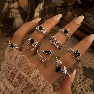 May include: A collection of silver-toned rings worn on fingers, featuring various designs including a bat with a black teardrop stone, a spider, a rose, thorny vines, and other intricate shapes with black accents and small white pearls.