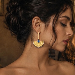May include: A gold-toned earring featuring a semi-circular fan shape with a textured surface. The earring is accented with a single teardrop-shaped blue stone at the top and a row of small white pearls lining the bottom edge of the fan.