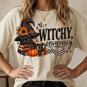 May include: A cream-colored t-shirt featuring a graphic of a black cat wearing a witch hat, perched on a stack of books next to a pumpkin and a mug. The text reads "WITCHY, OBVIOUSLY" in black lettering, accompanied by small stars and hearts.