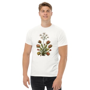 May include: A white short-sleeved t-shirt featuring a central graphic illustration of a Venus flytrap plant. The botanical design displays green leaves with red-tinted, spiky-edged traps and small white flowers extending from the top of the plant.