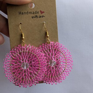 Puede incluir: Un par de pendientes colgantes redondos con cuentas de semillas de color rosa vibrante dispuestas en un patrón circular alrededor de una cuenta de cristal transparente central. Tienen ganchos de metal dorado y están en una tarjeta marrón Handmade with love.