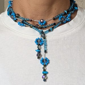 Puede incluir: Un collar de cuentas de varias hebras con cuentas de color azul brillante y oscuras iridiscentes. El diseño incluye pequeños grupos de cuentas en forma de flor y dos hebras colgantes que caen desde el centro, acentuadas con fragmentos de piedra turquesa.