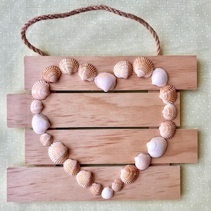 May include: A wall hanging sign made of four horizontal light brown wooden slats connected by a brown rope loop at the top. A heart shape is formed by twenty-four small, ridged, tan and white seashells arranged across the wooden surface.