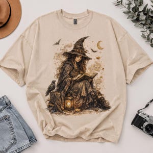 May include: A beige crewneck t-shirt featuring a detailed graphic print of a witch wearing a pointed hat, sitting on a stump while reading a book. The illustration includes a lantern, a raven, bats, and a crescent moon in sepia tones.