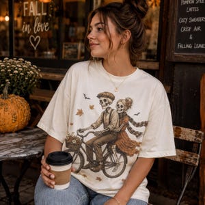 Puede incluir: Una camiseta de manga corta color hueso con una ilustración gráfica de dos esqueletos montando una bicicleta juntos. El esqueleto masculino lleva una gorra plana y pantalones, mientras que el esqueleto femenino lleva un vestido y una bufanda.