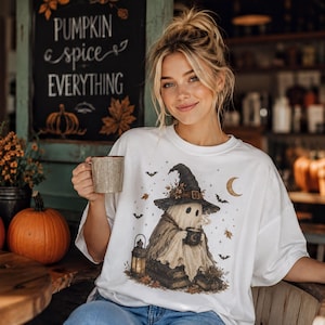 May include: A white t-shirt featuring a graphic of a ghost wearing a witch hat, holding a mug, and sitting near a lantern and books. The design includes autumn leaves, bats, and a crescent moon. The shirt is worn with blue denim jeans.