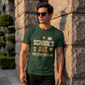 May include: A dark green short-sleeve t-shirt featuring the text "SCHOOL'S OUT. FREEDOM MODE ON." in a light-colored font. The graphic includes small icons of a document, a sun, an open book, a coffee cup, and a vinyl record.