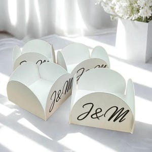 May include: A set of four white, curved, open-top paper favor boxes. Each box features the letters J&M printed in a black, elegant script font on the front panel. The boxes have a distinct scalloped edge design and a smooth, matte finish.
