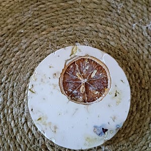 May include: A circular white coaster featuring an embedded dried orange slice in the center. The surface of the coaster is textured with small, scattered botanical elements, including tiny dried flower petals and leaves, creating a natural look.