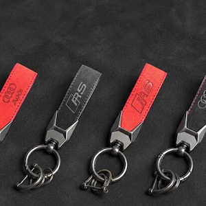 May include: Four keychains featuring a strap design with a metallic attachment and a circular ring. Two keychains are red with the Audi logo, and two are black, with one featuring the RS logo and the other featuring the Audi logo in a debossed style.
