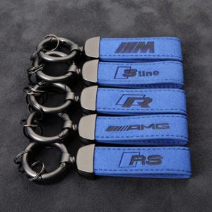 Op de afbeelding: Vijf blauwe suède sleutelhangers met donker metalen beslag en ronde ringen. Elke sleutelhanger heeft een ander in reliëf aangebracht logo: M, S line, R, AMG en RS. Het gestructureerde blauwe materiaal is langs de randen gestikt en verbonden met een metalen sluiting.