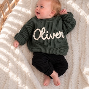 May include: A dark green long-sleeve ribbed knit jumper featuring the name Oliver embroidered in white yarn across the chest, paired with solid black ribbed knit trousers. The jumper has a crew neckline and cuffed sleeves, providing a textured look.