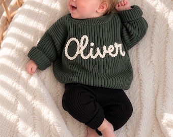 Puede incluir: Un suéter de manga larga en tejido de punto acanalado de color verde oscuro con el nombre Oliver bordado en hilo blanco en el pecho, combinado con pantalones de punto acanalado en color negro sólido. El suéter tiene cuello redondo y puños.