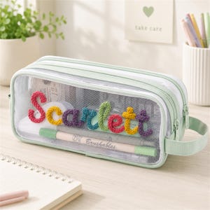 Puede incluir: Un estuche rectangular de malla transparente con bordes verde claro y cremallera a juego. En la parte frontal aparece el nombre Scarlett escrito con letras coloridas, texturizadas y similares a la lana. Incluye un asa lateral verde claro.