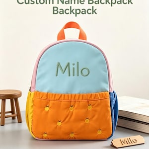 May include: A multi-coloured backpack featuring a light blue upper section with the embroidered name Milo, an orange quilted lower pocket with small embroidered orange fruit motifs, a pink trim, and an orange top handle. The text Custom Name Backpack Backpack is visible above, and the name Milo is on a wooden tag.