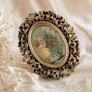 May include: An oval picture frame with an ornate, dark gold-toned metallic border featuring intricate scrollwork and leaf motifs. The frame contains a print of a child in a light-colored dress standing near a rose bush with pink flowers.