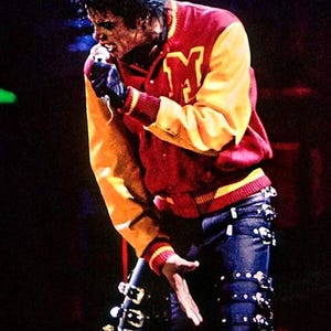 May include: A red and yellow varsity jacket with a letter M on the chest, worn with dark blue trousers featuring multiple black straps and silver buckles. The attire includes a single black glove on one hand while holding a microphone near the mouth.