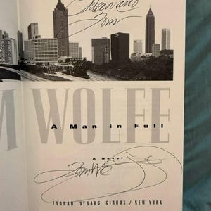 May include: A title page from the book A Man in Full by Tom Wolfe, featuring a black and white photograph of the Atlanta skyline at the top, the author's name and book title in black text, and two distinct handwritten ink signatures across the page.
