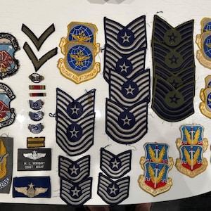 May include: A collection of embroidered military patches and rank insignia, including Air Force Reserve and Military Airlift Command emblems, chevron stripes, and a black name tag reading H. L. WRIGHT SSGT USAF, featuring various blue, gold, and white designs.
