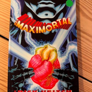 May include: A graphic novel titled The Maximortal by Rick Veitch, volume 1 of The King Hell Heroica. The cover features a large, anatomical human heart held by two dark, gloved hands against a blue, lightning-filled background.