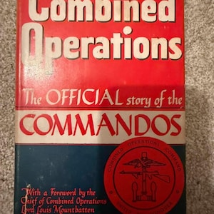 May include: A hardcover book titled Combined Operations: The OFFICIAL story of the COMMANDOS. The cover features a red top section with white text, a blue bottom section, and a circular red emblem depicting a weapon and anchor symbol.