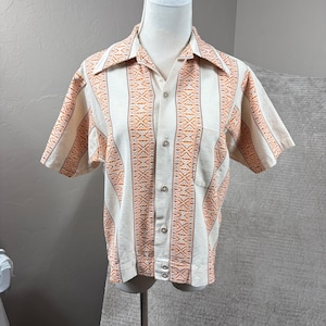 May include: A short-sleeve button-down shirt in an off-white color featuring vertical stripes with an intricate orange and brown geometric pattern. The shirt has a pointed collar, a single chest pocket on the left side, and light-colored buttons.