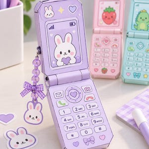 May include: A purple paper craft flip phone featuring a screen with a white bunny illustration, a keypad with alphanumeric buttons, and a purple beaded charm with a matching bunny pendant. Two additional paper flip phones in pink and blue are visible.
