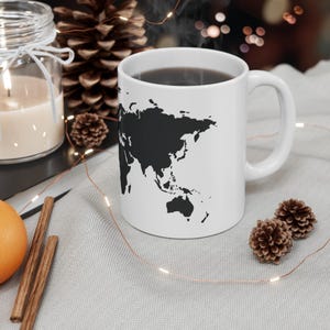 May include: A white ceramic mug featuring a black silhouette of a world map printed on the front. The mug has a standard cylindrical shape with a curved handle on the right side and is filled with a dark liquid.