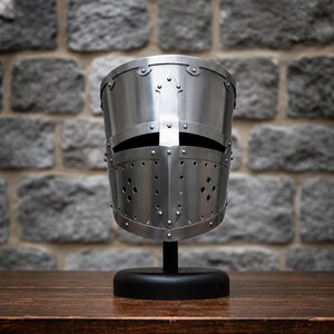 May include: A polished steel great helm with a flat top and a cross-shaped faceplate featuring a horizontal eye slit and circular ventilation holes, mounted on a black circular stand. The helmet is constructed with visible metal rivets.