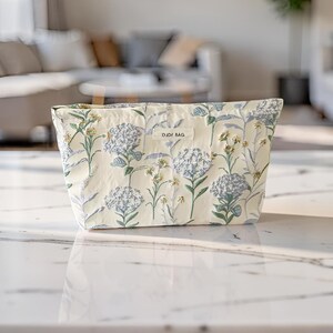 May include: A cream-coloured pouch featuring a botanical print of blue hydrangea flowers and small yellow wildflowers. The bag has a rectangular shape and a central fabric label with the text DJDF BAG printed in black on the front surface.